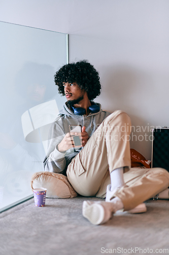 Image of African-American entrepreneur taking a relaxing break from work, sitting on the floor while using wireless headphones and a smartphone for some digital entertainment.