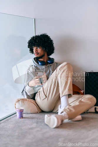 Image of African-American entrepreneur taking a relaxing break from work, sitting on the floor while using wireless headphones and a smartphone for some digital entertainment.