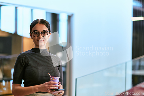 Image of In a bustling modern office a businesswoman in glasses juggles her tasks, sipping coffee and using her smartphone, epitomizing the dynamic and multitasking nature of contemporary corporate life