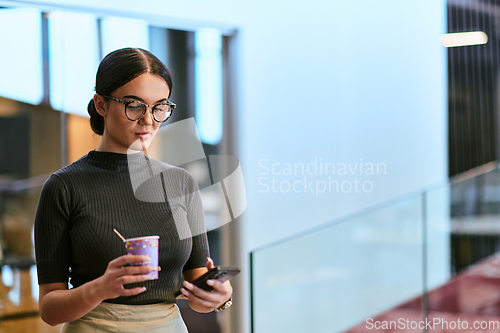 Image of In a bustling modern office a businesswoman in glasses juggles her tasks, sipping coffee and using her smartphone, epitomizing the dynamic and multitasking nature of contemporary corporate life