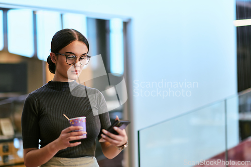 Image of In a bustling modern office a businesswoman in glasses juggles her tasks, sipping coffee and using her smartphone, epitomizing the dynamic and multitasking nature of contemporary corporate life