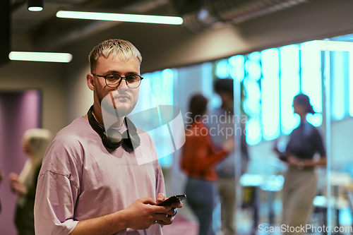 Image of In a contemporary startup office, a modern blond entrepreneur stands, engrossed in his smartphone, epitomizing the dynamic and tech-savvy essence of the modern business world.