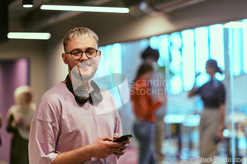 Image of In a contemporary startup office, a modern blond entrepreneur stands, engrossed in his smartphone, epitomizing the dynamic and tech-savvy essence of the modern business world.