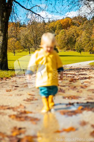 Image of Sun always shines after the rain. Small bond infant boy wearing yellow rubber boots and yellow waterproof raincoat walking in puddles in city park on sunny rainy day.