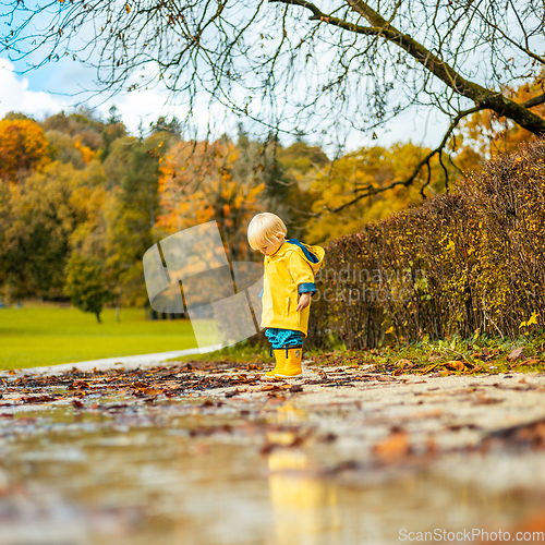 Image of Sun always shines after the rain. Small bond infant boy wearing yellow rubber boots and yellow waterproof raincoat walking in puddles in city park on sunny rainy day.