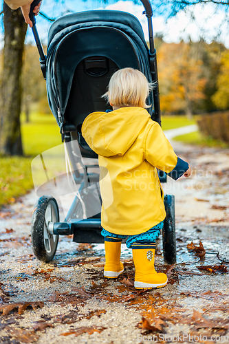 Image of Sun always shines after the rain. Small blond infant boy wearing yellow rubber boots and yellow waterproof raincoat walking in puddles, pushing stroller in city park, holding mother's hand after rain.