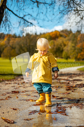 Image of Sun always shines after the rain. Small bond infant boy wearing yellow rubber boots and yellow waterproof raincoat walking in puddles in city park on sunny rainy day.