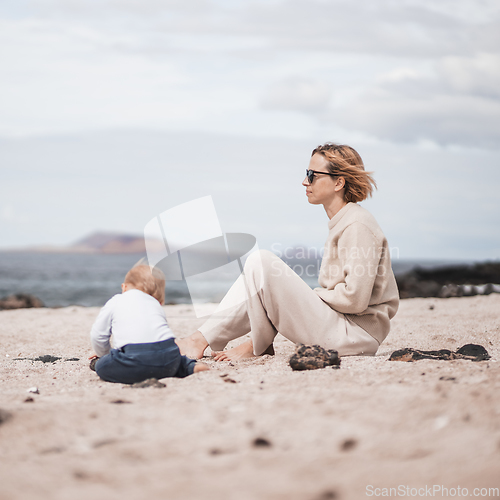 Image of Mother enjoying winter beach vacations playing with his infant baby boy son on wild volcanic sandy beach on Lanzarote island, Canary Islands, Spain. Family travel and vacations concept
