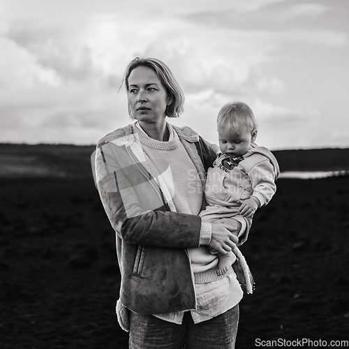 Image of Mother enjoying winter vacations playing with his infant baby boy son on black sandy volcanic beach of Janubio on Lanzarote island, Spain on windy overcast day. Family travel vacations concept.