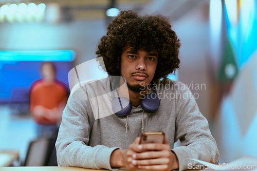 Image of In a modern startup office, an African American man, balancing work and technology, utilizes headphones and a smartphone, emblematic of contemporary multitasking and productivity