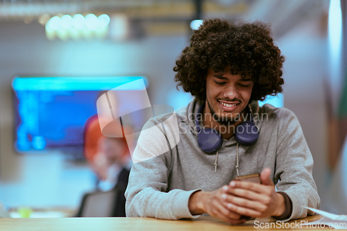Image of In a modern startup office, an African American man, balancing work and technology, utilizes headphones and a smartphone, emblematic of contemporary multitasking and productivity
