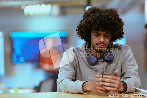 Image of In a modern startup office, an African American man, balancing work and technology, utilizes headphones and a smartphone, emblematic of contemporary multitasking and productivity