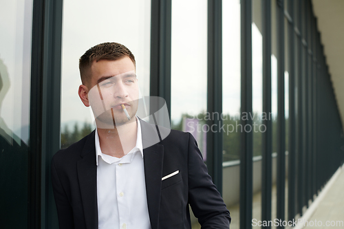 Image of Amidst the corporate hustle, a modern businessman in a black suit takes a smoke break outside his workplace, seeking a moment of relaxation in the midst of a busy day.