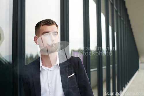 Image of Amidst the corporate hustle, a modern businessman in a black suit takes a smoke break outside his workplace, seeking a moment of relaxation in the midst of a busy day.