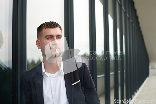 Image of Amidst the corporate hustle, a modern businessman in a black suit takes a smoke break outside his workplace, seeking a moment of relaxation in the midst of a busy day.