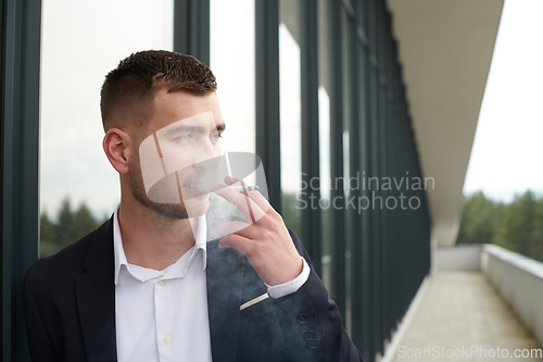 Image of Amidst the corporate hustle, a modern businessman in a black suit takes a smoke break outside his workplace, seeking a moment of relaxation in the midst of a busy day.