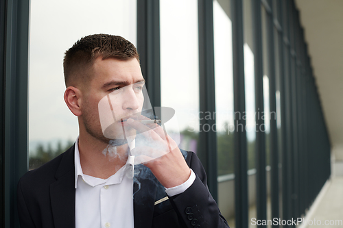 Image of Amidst the corporate hustle, a modern businessman in a black suit takes a smoke break outside his workplace, seeking a moment of relaxation in the midst of a busy day.