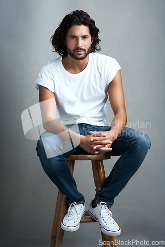 Image of Fashion, handsome and portrait of man in studio on a stool with casual, cool and stylish outfit. Confidence, serious and young male model from Mexico with trendy style on chair by gray background.