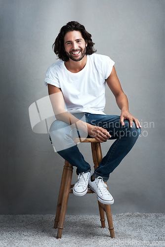 Image of Fashion, happy and portrait of man in studio on a stool with casual, cool and stylish outfit. Smile, handsome and confident young male model from Mexico with trendy style on chair by gray background.