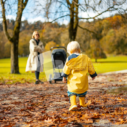 Image of Sun always shines after the rain. Small bond infant boy wearing yellow rubber boots and yellow waterproof raincoat walking in puddles and autumn leaves in city park on sunny rainy day.