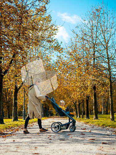 Image of Young beautiful mother wearing a rain coat pushing stroller with her little baby boy child, walking in city park on a sunny autumn day