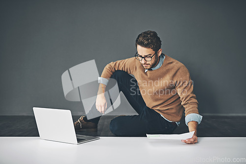 Image of Business man working on a laptop, reading a report and going through finance paperwork while sitting on the floor in an office at work. Serious professional manager looking at papers with copyspace