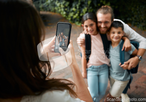 Image of Mother taking a family photo on her phone of her happy little children and their father smiling before school. Boy and girl siblings with their dad smile for a picture. Mom taking photos of kids