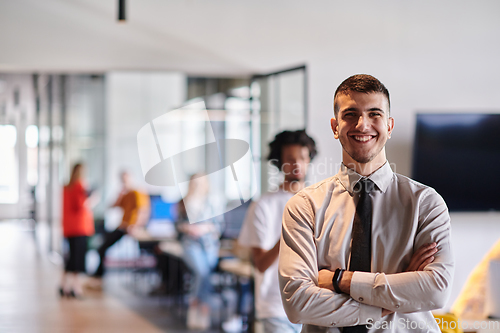 Image of A young business leader stands with crossed arms in a modern office hallway, radiating confidence and a sense of purpose, embodying a dynamic and inspirational presence.