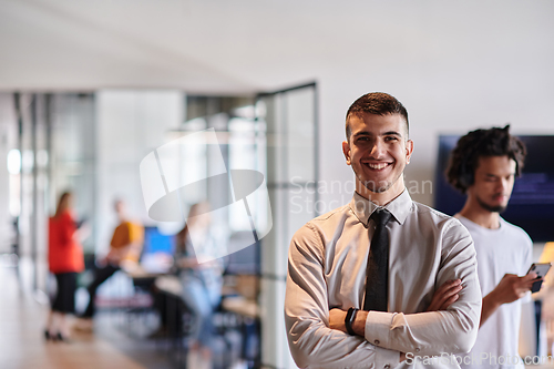 Image of A young business leader stands with crossed arms in a modern office hallway, radiating confidence and a sense of purpose, embodying a dynamic and inspirational presence.