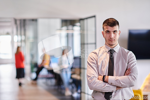 Image of A young business leader stands with crossed arms in a modern office hallway, radiating confidence and a sense of purpose, embodying a dynamic and inspirational presence.