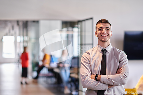 Image of A young business leader stands with crossed arms in a modern office hallway, radiating confidence and a sense of purpose, embodying a dynamic and inspirational presence.