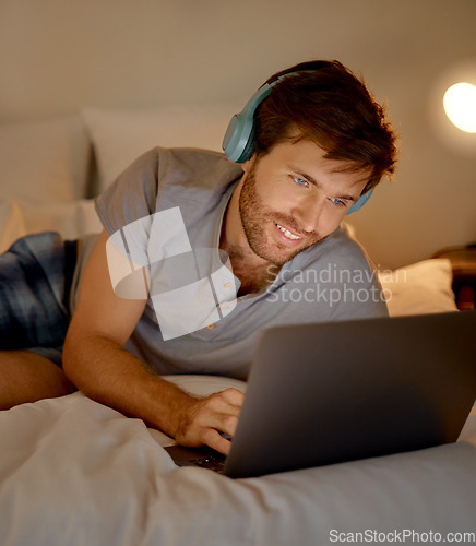 Image of Typing, headphones and in bed on laptop young man looking for movie or video to watch and relax at night in home. Happy male on internet and listening to music on streaming website and social media