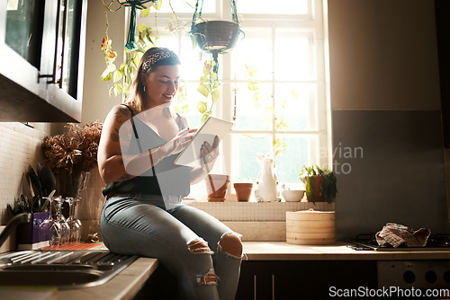 Image of Trendy woman browsing internet on tablet using her new wifi at home looking happy and satisfied. Normal, real and edgy young female searching online for cool recipe ideas to use in her modern kitchen