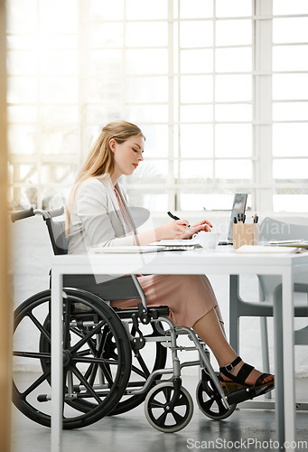 Image of Professional, disabled business woman in wheelchair reading documents, writing or making notes on office desk sitting by laptop. Female entrepreneur with disability doing contract paperwork with pen.