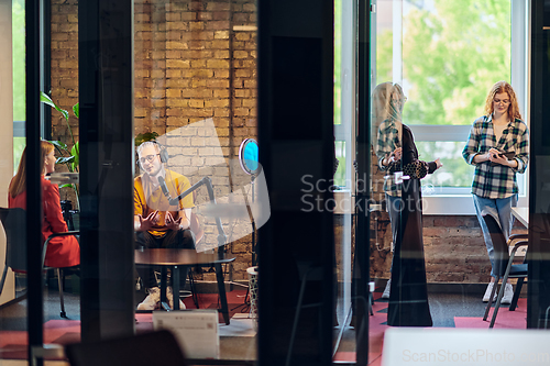 Image of A gathering of young business professionals, some seated in a glass-walled office, engage in a lively conversation and record an online podcast, embodying modern collaboration and dynamic interaction