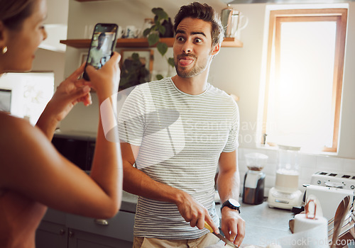 Image of Silly, goofy and interracial couple taking pictures, having fun and cooking together in the kitchen at home. Happy girlfriend taking pictures of boyfriend, making funny faces and preparing dinner