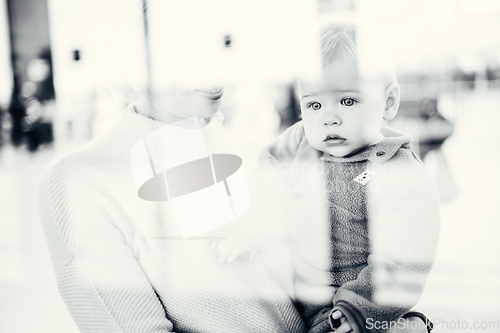 Image of Thoughtful young mother looking trough window holding his infant baby boy child while waiting to board an airplane at airport terminal departure gates. Travel with baby concept.