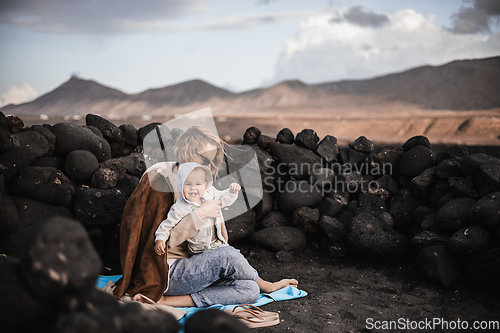 Image of Mother enjoying winter vacations playing with his infant baby boy son on black sandy volcanic beach of Janubio on Lanzarote island, Spain on windy overcast day. Family travel vacations concept.