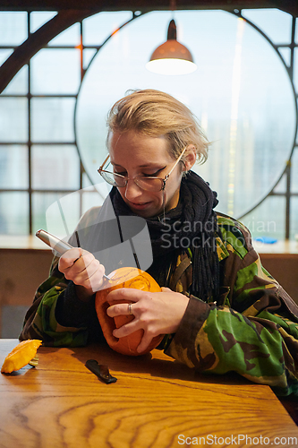 Image of A modern blonde woman in military uniform is carving spooky pumpkins with a knife for Halloween night