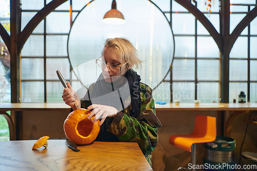 Image of A modern blonde woman in military uniform is carving spooky pumpkins with a knife for Halloween night