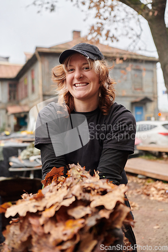 Image of A stylish, modern young woman takes on the role of a garden caretaker, diligently collecting old, dry leaves and cleaning up the yard in an eco-conscious manner