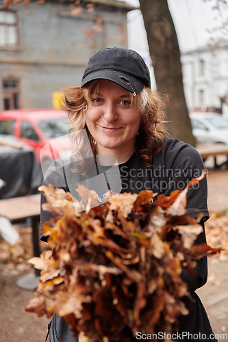 Image of A stylish, modern young woman takes on the role of a garden caretaker, diligently collecting old, dry leaves and cleaning up the yard in an eco-conscious manner