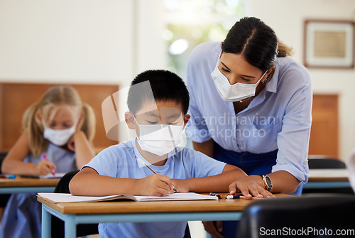 Image of Covid, education and learning with a teacher wearing a mask and helping a male student in class during school. Young boy studying in a classroom with help from an educator while sitting at his desk.