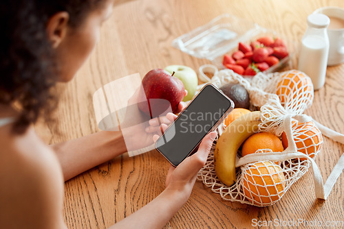 Image of Colorful, healthy and nutritional organic fruit groceries on table beside woman holding phone and looking online for smoothie recipes. Young lady eating fresh organic apple for wellness vegan diet