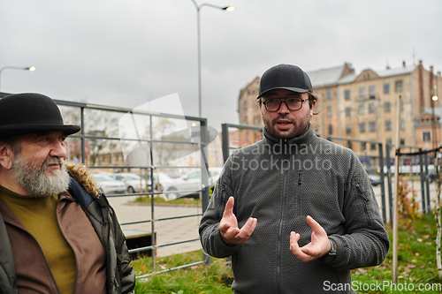 Image of In the picturesque garden of Riga, Latvia, a man engages in a heartfelt conversation with his older friend, enjoying the serene urban oasis amidst the bustling cityscape