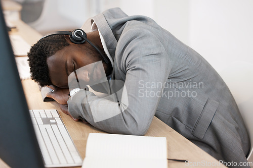 Image of Tired CRM call center agent sleeping on computer desk at work office with headset on. Exhausted young business man taking a nap on table. African American male feeling burnout resting in workplace.