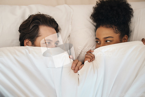 Image of Interracial, happy and relaxed couple lying in bed, bonding and looking shy while hiding after waking up together in the morning. Loving, carefree and romantic boyfriend and girlfriend being intimate