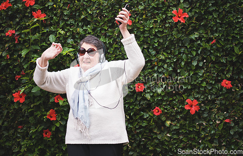 Image of Funny old woman dancing listening to music on smartphone wearing earphones smiling enjoying fun celebrating retirement in garden with flower wall