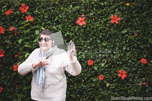 Image of Funny old woman dancing listening to music on smartphone wearing earphones smiling enjoying fun celebrating retirement in garden with flower wall