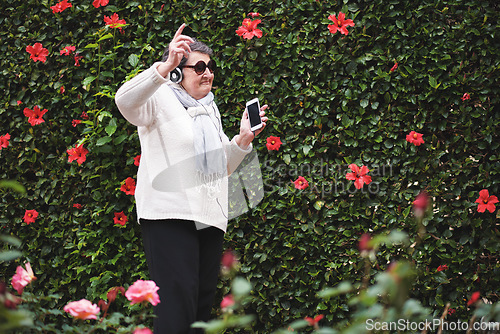 Image of Funny old woman dancing listening to music on smartphone wearing earphones smiling enjoying fun celebrating retirement in garden with flower wall